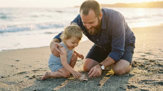 Bearded father kneeling beside his toddler at a sandy beach, both examining a seashell together, father's protective arm around child's shoulders, child reaching toward