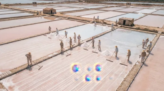 Close aerial view of a traditional salt harvesting operation on coastal flats, workers in wide-brimmed hats raking crystallized salt into low white pyramids along flooded evaporation channels, the