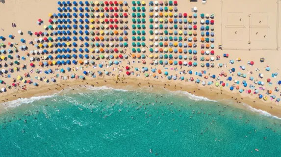 An aerial view of a packed summer beach with brightly colored umbrellas creating a mosaic pattern across the sand, clear turquoise shallow water at the bottom of the frame, tiny figures wading in the