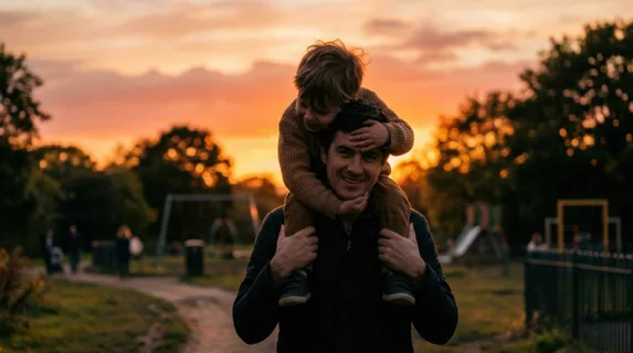 Young child sitting on an adult's shoulders at sunset in a park setting, child's arms wrapped around head in affectionate embrace, both figures silhouetted against