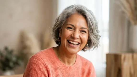 Mature woman with grey highlighted hair and radiant genuine smile, warm-toned skin catching diffused studio light, subtle laugh lines framing mouth and eyes showing