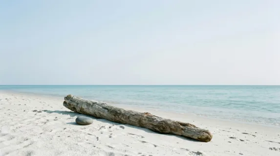 Lone driftwood log resting diagonally on pristine white sand near calm turquoise water, single smooth stone placed beside it, cloudless pale sky dominating the