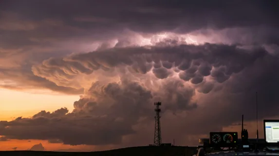 Cumulonimbus anvil cloud lit from within by continuous lightning at golden hour, cell tower silhouette in foreground, mammatus pouches hanging from cloud base like inverted cobblestones, storm
