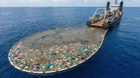 An ocean plastic collection vessel at sea, large booms gathering surface debris, the contrast between the clean blue water and the collected waste vivid and stark, the scale of the problem and the