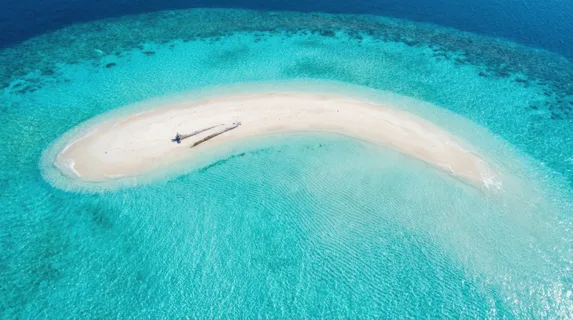 A pristine Maldivian sandbank at high tide, the sand a narrow white arc surrounded on all sides by turquoise water grading to deep cobalt at the reef edge, overwater bungalows absent, only a single