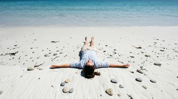 Overhead view of a person lying flat on pristine white sand with arms stretched outward, wearing casual beach attire, surrounded by scattered smooth pebbles and seaweed,