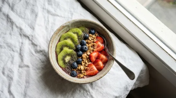 A just-made smoothie bowl topped with sliced kiwi, strawberries, blueberries, granola and chia seeds, the bowl placed on a white linen cloth by a window, morning light catching the dewy surface of the