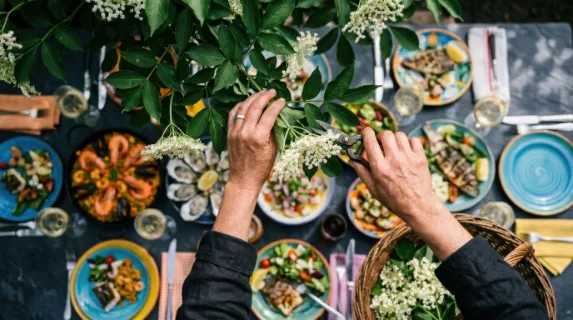 an overhead angle of hands harvesting elderflowers, framed against the vibrant backdrop of a blurred array of colorful seafood dishes on a slate surface. the scene