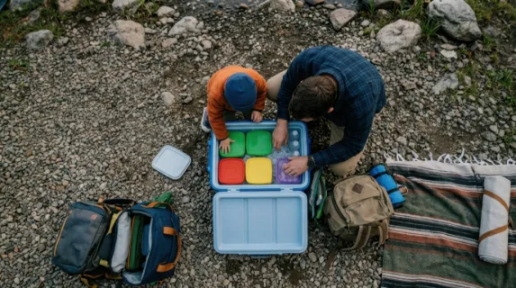 an overhead shot of the father and child crouching together, packing the cooler with vibrant containers, the serene lake visible in the blurred background, creating an