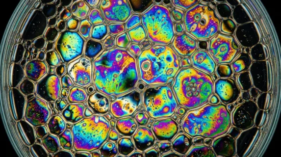 Abstract macro of oil and water emulsion in a shallow dish under polarized light, psychedelic interference colors blooming across irregular bubble formations, each oil