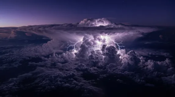 A thunderstorm cell viewed from above the clouds, the tops of cumulonimbus towers lit by lightning from within, electric veins branching across the cloud surface in