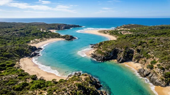 Aerial view of a winding coastal inlet with turquoise water cutting through sandy shores and rocky edges, gentle waves forming patterns along the coastline, scattered