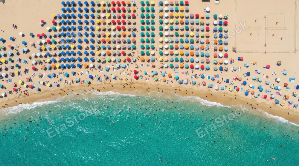 An aerial view of a packed summer beach with brightly colored umbrellas creating a mosaic pattern across the sand, clear turquoise shallow water at the bottom of the frame, tiny figures wading in the