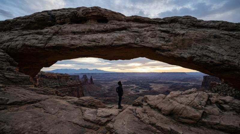 A person standing under an arch of rocks, rugged textures, vast landscape, small human scale against nature, contemplative atmosphere, ultra-realistic, no logos.