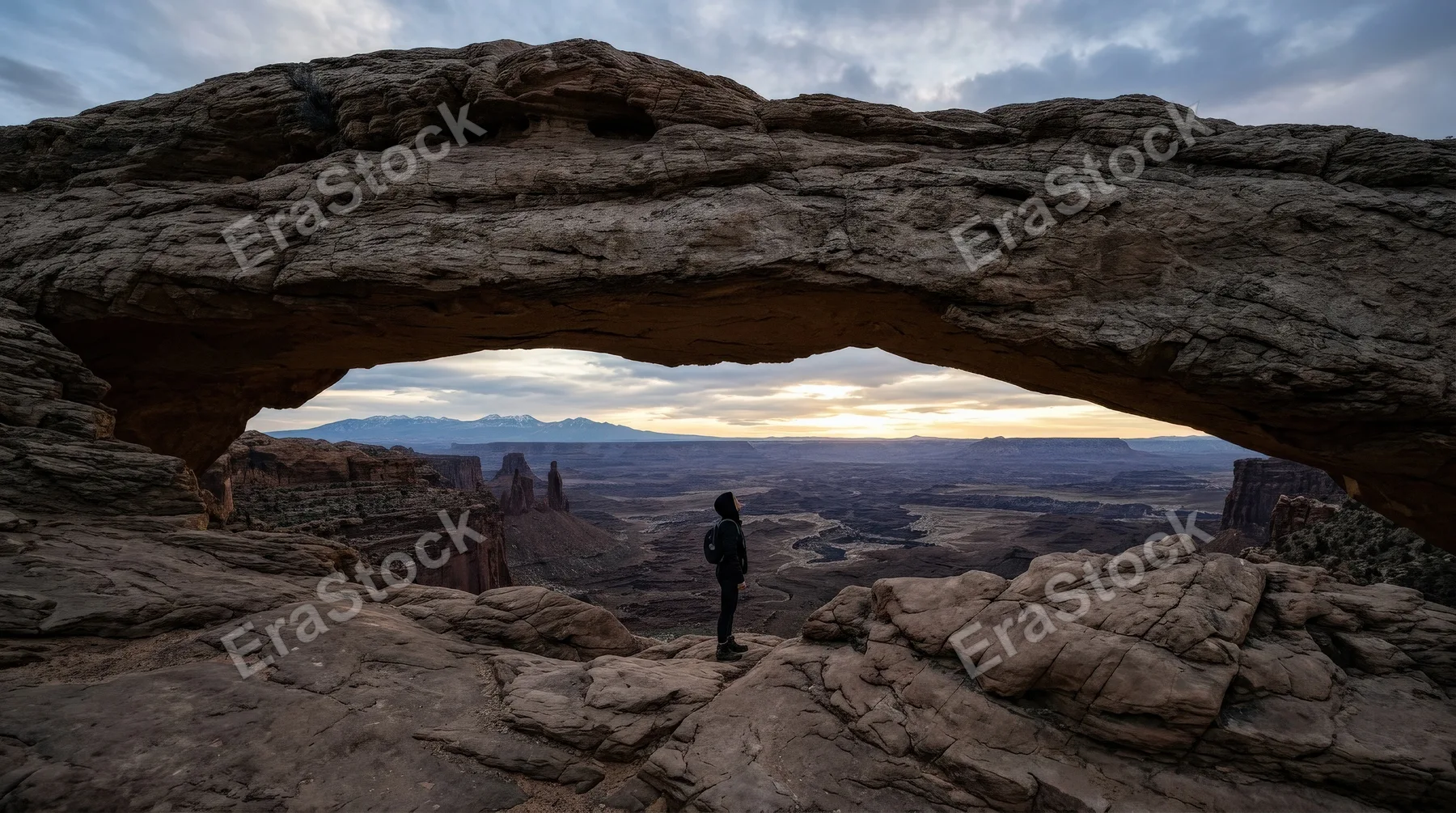 A person standing under an arch of rocks, rugged textures, vast landscape, small human scale against nature, contemplative atmosphere, ultra-realistic, no logos.