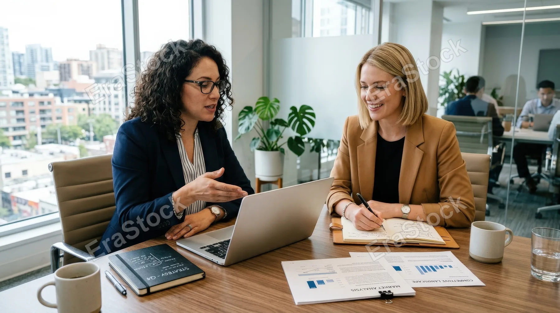 Two businesswomen discussing ideas using a laptop and taking notes in an office, seated side by side at a sleek table, documents and pens adding realistic detail, bright workspace and a focused