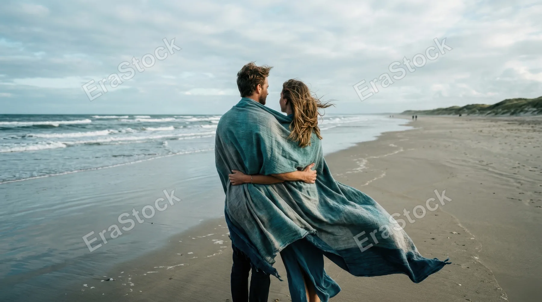 Couple standing on a wide open beach wrapped in a large flag fabric, arms around each other, looking toward the horizon where sea meets sky, fabric flowing dramatically