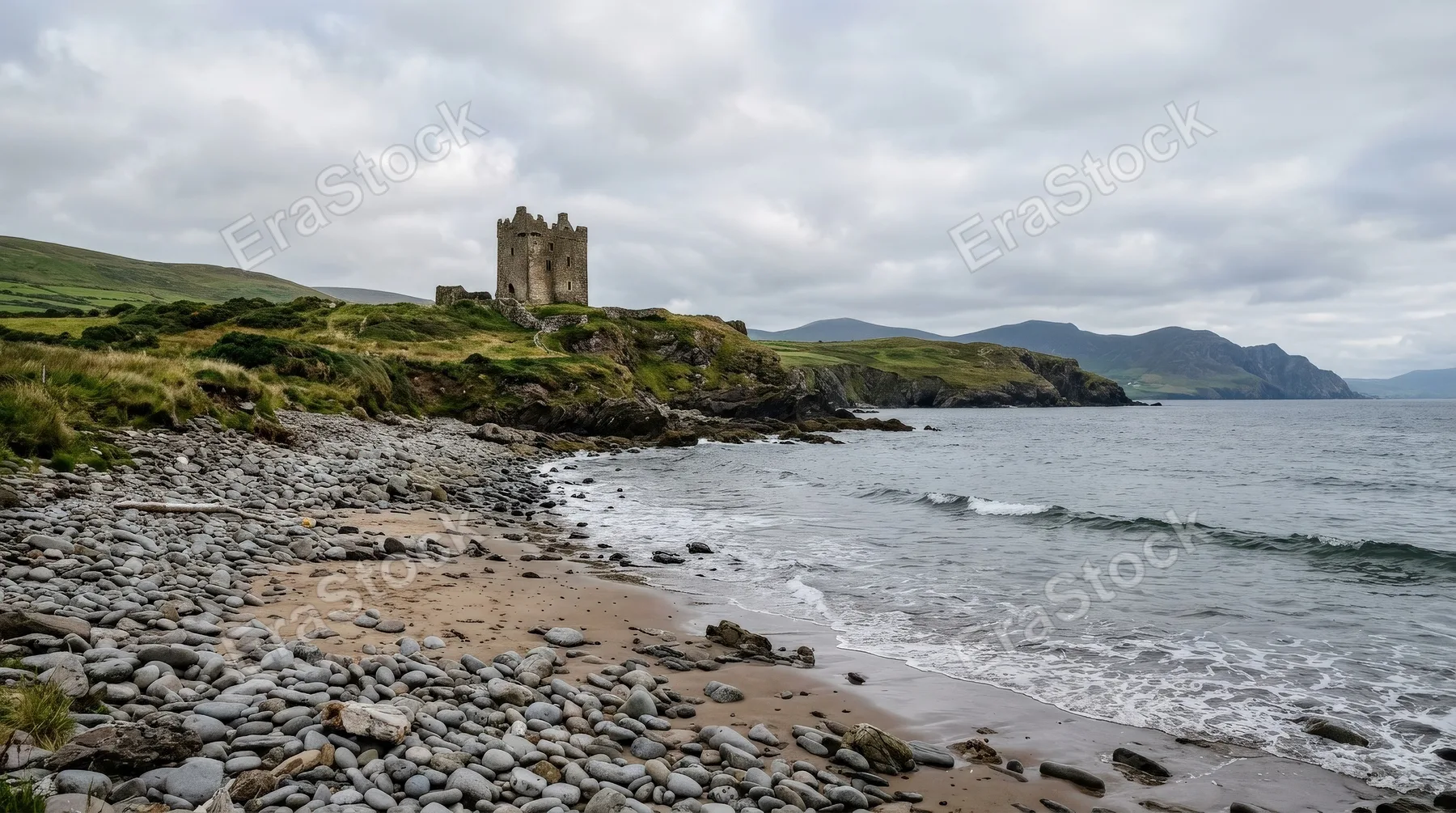 A sandy beach strewn with smooth stones and Minard Castle rising in the background on the Dingle Peninsula, windswept Irish coastline under soft overcast light, rugged textures and moody Atlantic