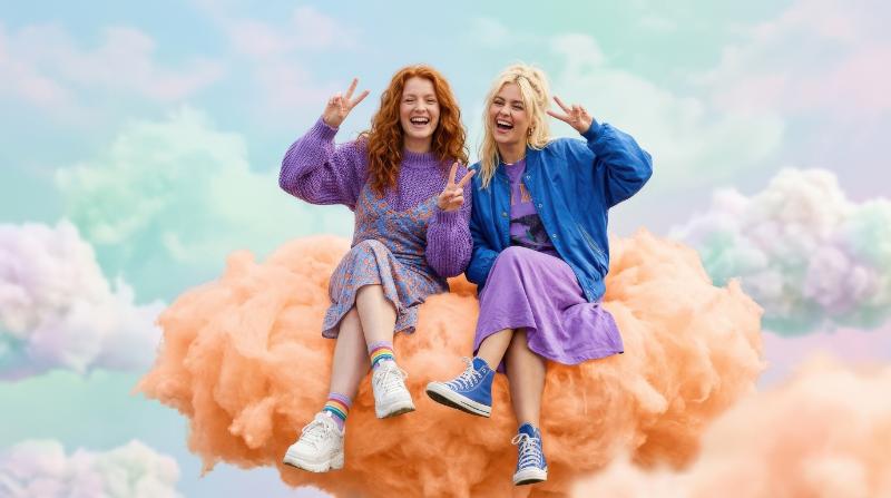 Two funny colorful young women with red and blonde hair, fashion-forward blue and purple outfits, sitting on an orange cloud and flashing peace signs, airy pastel setting with joyful carefree vibe,
