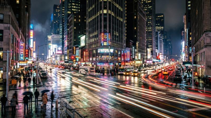 A busy city intersection at night captured in dynamic motion, glowing traffic streams, neon reflections on wet asphalt, towering buildings, electric urban energy, fast-paced metropolitan atmosphere,