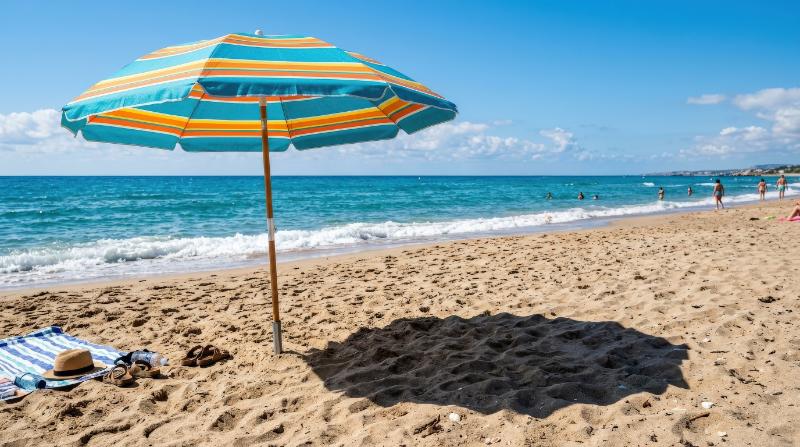 A colorful beach umbrella casting a sharp shadow on the sand with the ocean beyond, bright sunlight and vivid summer contrast, ultra-realistic, no logos.