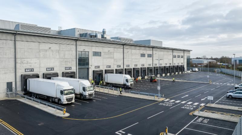 A modern industrial warehouse exterior with multiple loading bays, clean concrete facade, delivery docks, wide asphalt yard, functional logistics architecture, cool daylight, efficient professional