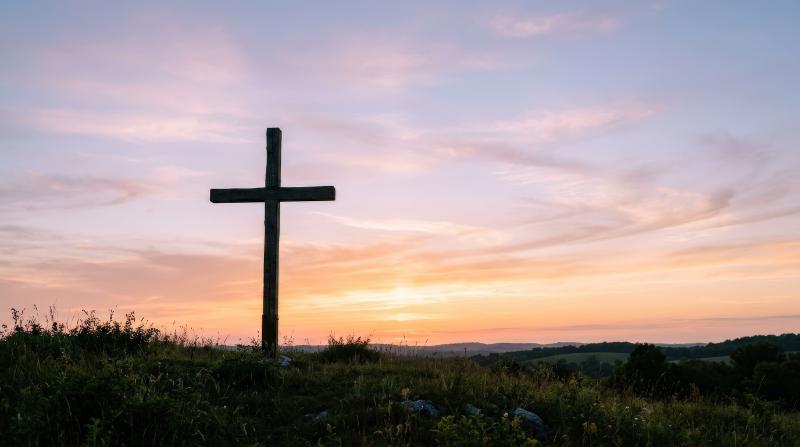 A wooden Christian cross silhouette against a soft pastel sunset sky, minimal composition, tranquil horizon glow and peaceful spiritual atmosphere, ultra-realistic, no logos.