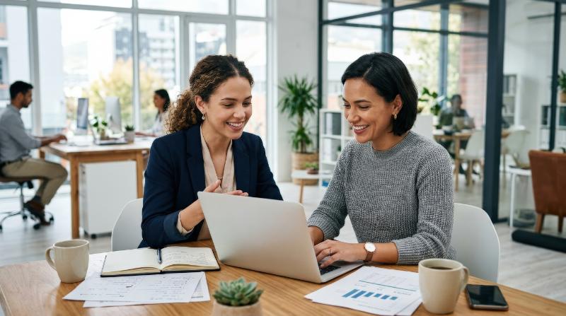 Two female professionals working together on a laptop in a bright office environment, side-by-side at a shared desk, papers nearby, airy daylight, positive collaborative atmosphere, ultra-realistic,